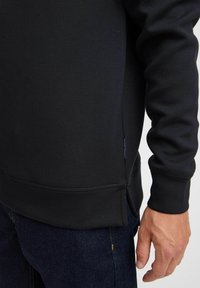 Sweatshirt noir avec une texture lisse, détails à fente sur les côtés et une étiquette de marque visible au niveau de l'ourlet. Associé à un jeans en denim foncé.