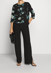 Sort blomsterbluse med teal og hvide accenter, trekvartærmer; parret med sorte brede bukser med sideslids og sorte sandaler.