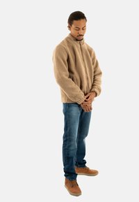 Uomo con capelli intrecciati che indossa una giacca in pile marrone chiaro, jeans blu e scarpe marroni, in piedi con le mani unite e la testa leggermente chinata.