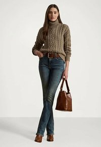 Nő barna kötött garbó pulóverben, sötétkék farmernadrágban, barna övvel, barna bokacsizmában, egy barna textúrált táskával a kezében.