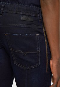 Jeans de mezclilla azul oscuro con un bolsillo trasero que presenta un costurón amarillo en contraste y una pequeña etiqueta negra. Detalle notable de cremallera lateral.