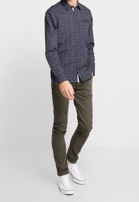 Hombre con camisa de manga larga azul marino con estampado, pantalones ajustados color verde oliva y zapatillas blancas de caña alta, de pie con una mano en el bolsillo.