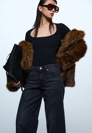 Sieviete valkā lielizmēra saulesbrilles, melnu garām piedurknēm džemperi, tumšas plaša piegriezuma džinsas, rokās tur brūnu mākslīgās kažokādas mēteļa un melnu teksturētu pleca somu.