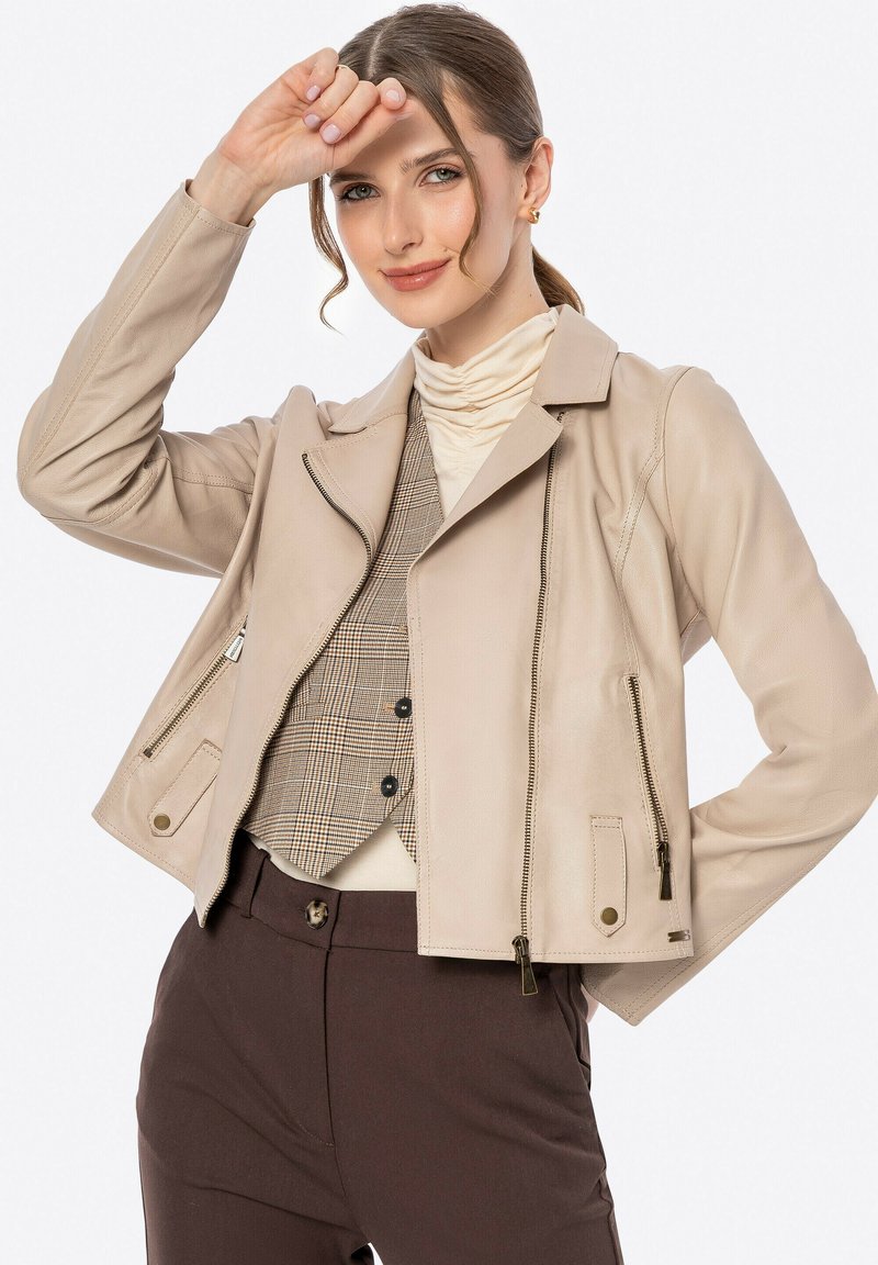 Beige leren motorjack met een gekartelde kraag, ritssluiting aan de voorkant en zijzakken. Draag het over een beige coltrui en een geruite vest.