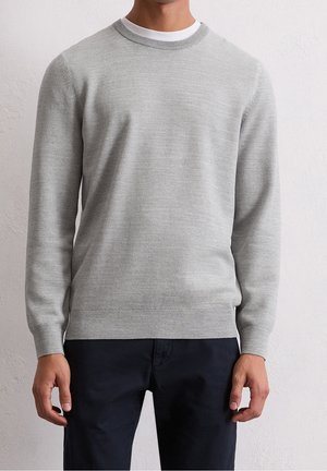 Homme portant un pull en maille texturée gris clair par-dessus un t-shirt blanc à col rond et un pantalon sombre, debout contre un mur clair uni.