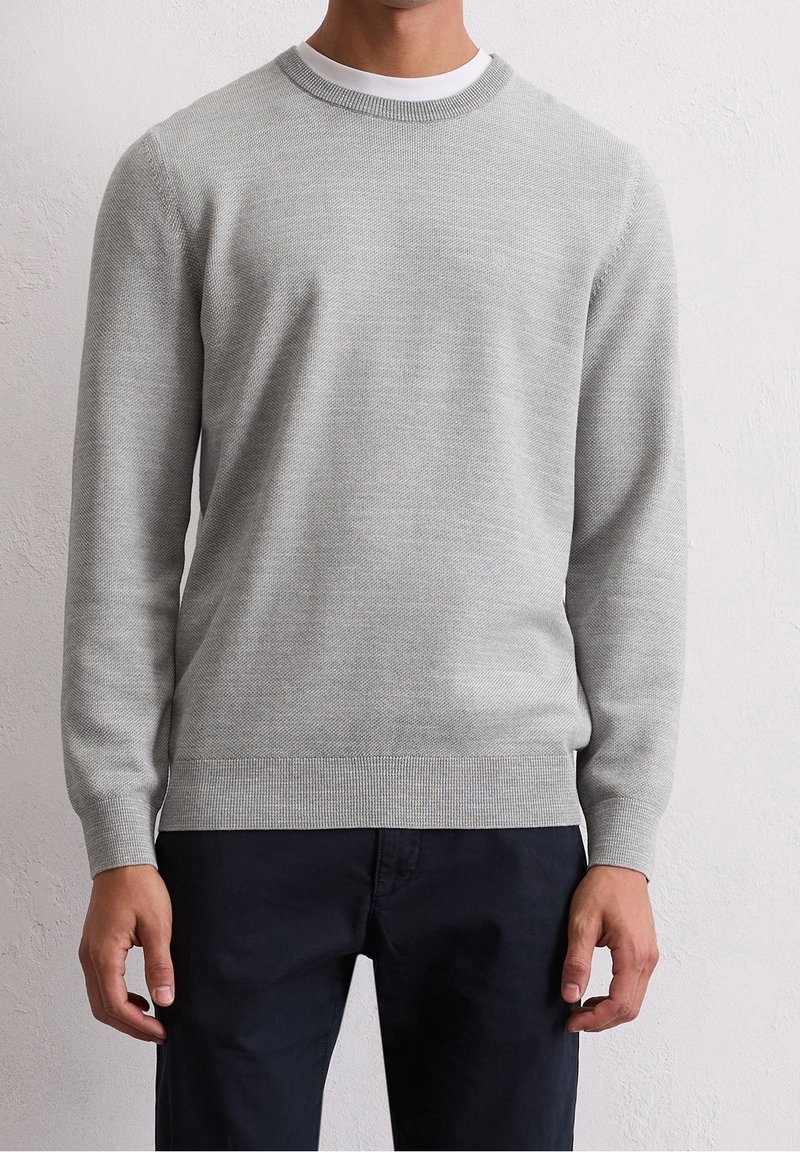 Homme portant un pull en maille texturée gris clair par-dessus un t-shirt blanc à col rond et un pantalon sombre, debout contre un mur clair uni.
