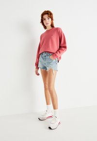 Lyserød sweatshirt, lyseblå slidte denimshorts, hvide sneakers med røde accenter, og hvide sokker, præsenteret mod en ensfarvet baggrund.