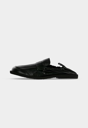 Chaussure noire en cuir sans talon, à enfiler, avec un bout arrondi et des côtés élastiques extensibles, présentée sur un fond uni gris clair.