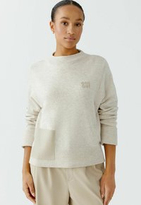 Hellbeiger Pullover mit geripptem Saum und Bündchen, der eine strukturierte aufgesetzte Tasche aus einem weichen, kontrastierenden Material aufweist. Dezentes Logodetail auf der Brust.