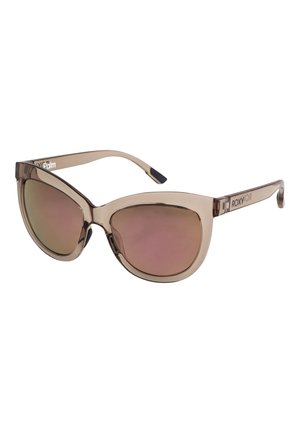 Des lunettes de soleil en œil de chat avec des montures transparentes couleur beige et des verres dégradés rose-brun. Elles présentent une finition brillante et un logo sur la branche.