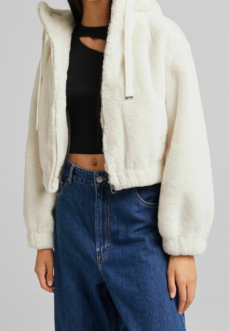 Klippt vit fälljacka med huva, dragkedjestängning och dragsnodd. Matchas med en svart ribbad topp och högmidjade blå denimjeans.