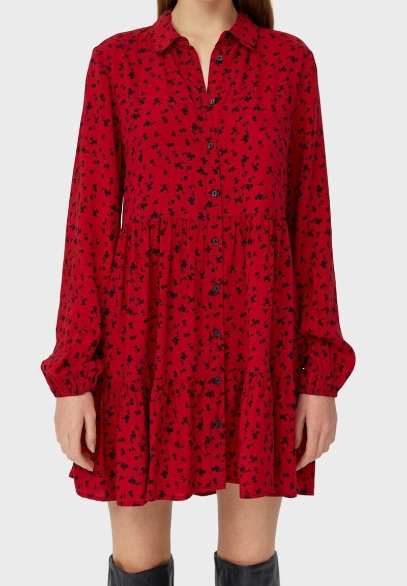 Femme portant une robe rouge à manches longues boutonnée avec de petits motifs floraux noirs et une jupe à volants, associée à des bottes noires montant jusqu’aux genoux.