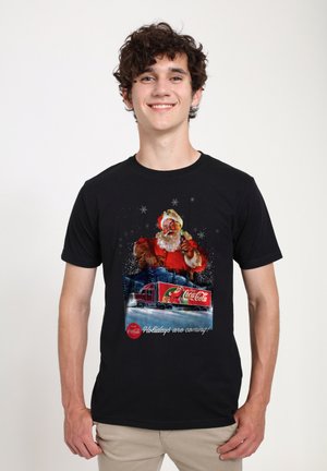 Black t-shirt featuring a Coca-Cola holiday graphic, with Santa Claus and a truck on a snowy background, accented by stars. Text reads, "Holidays are coming!"