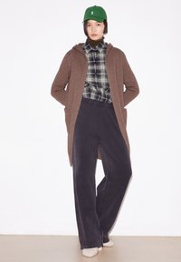 Brauner, langer Cardigan mit Taschen über einem schwarz-weißen karierten Hemd, kombiniert mit weiten, dunklen Hosen und einer grünen Mütze. Weiche Textur insgesamt.