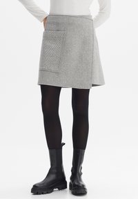 Grauer Tweed-Wickelrock mit diagonaler Saumlinie und großer aufgesetzter Tasche, kombiniert mit schwarzen Strumpfhosen und groben schwarzen Stiefeletten.