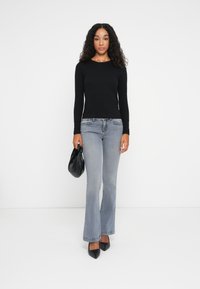 Schwarzes langärmliges Oberteil, hellgraue schmal geschnittene Jeans, schwarze spitze Absätze und eine kleine schwarze Tasche; glattes Material, eng anliegende Passform und detaillierte Nähte.
