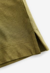 Olijfgroene katoenen stof met een vlakke naad, voorzien van een zijspleet aan de onderkant voor meer bewegingsvrijheid en een gladde textuur.
