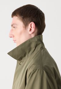 Olive green jacket with a high, folded collar, featuring a smooth texture and minimal stitching detail, shown from the side.