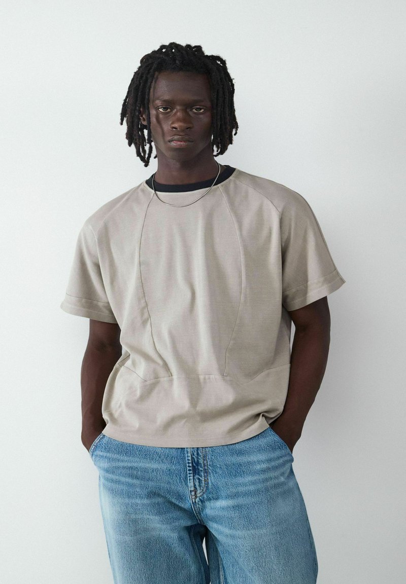 Beige T-shirt met korte mouwen en een relaxte pasvorm, voorzien van een asymmetrisch naadontwerp en een ronde halslijn, gedragen met lichtblauwe jeans.