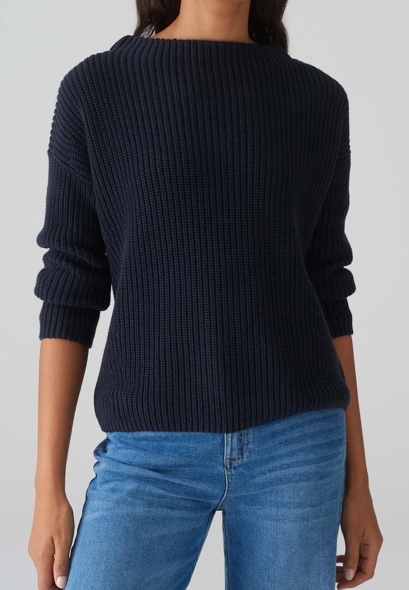 Pull en maille bleu marine avec une texture côtelée, large encolure bateau et manches retroussées, associé à un jean bleu clair.