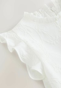 Tessuto bianco strutturato con maniche corte arricciate e collo alto arricciato, con una cerniera sul retro del collo.