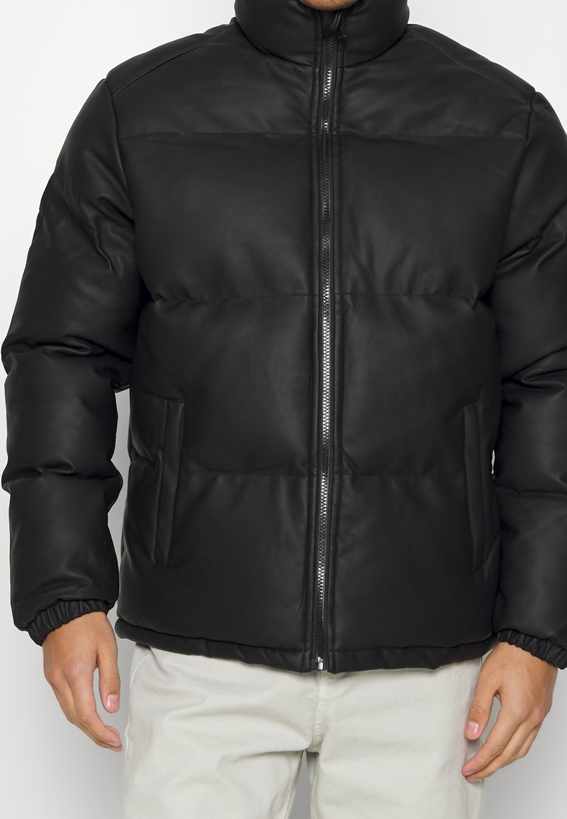 Mężczyzna w czarnej, pikowanej kurtce z przednim zamkiem błyskawicznym, wysokim kołnierzem, bocznymi kieszeniami i elastycznymi mankietami, w połączeniu z jasnymi spodniami.