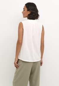 Blouse blanche sans manches avec un motif côtelé texturé et un ourlet arrondi. Le dos présente un léger froncé au niveau du col.