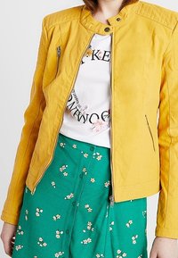 Giacca di pelle gialla con colletto alla mandarino e tasche con zip. Indossata sopra una T-shirt bianca e una gonna verde con un motivo floreale.