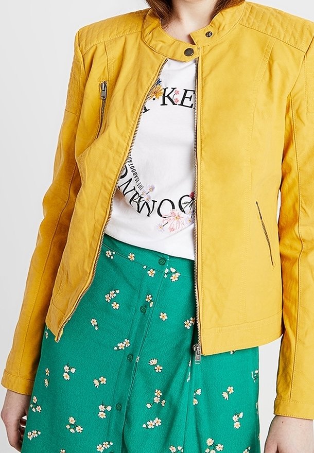 Giacca di pelle gialla con colletto alla mandarino e tasche con zip. Indossata sopra una T-shirt bianca e una gonna verde con un motivo floreale.