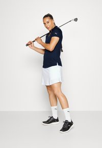 Tengerészkék poloing, fehér mintás szoknya, fekete golfcipő és fehér zoknik. Golf ütő a váll felett, atletikus póz.