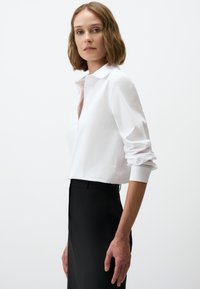 Weißes Blusenshirt mit umgeschlagenen Ärmeln, das eine glatte Textur aufweist. Kombiniert mit einem hoch taillierten, schwarzen Rock, der eine maßgeschneiderte Passform zeigt.