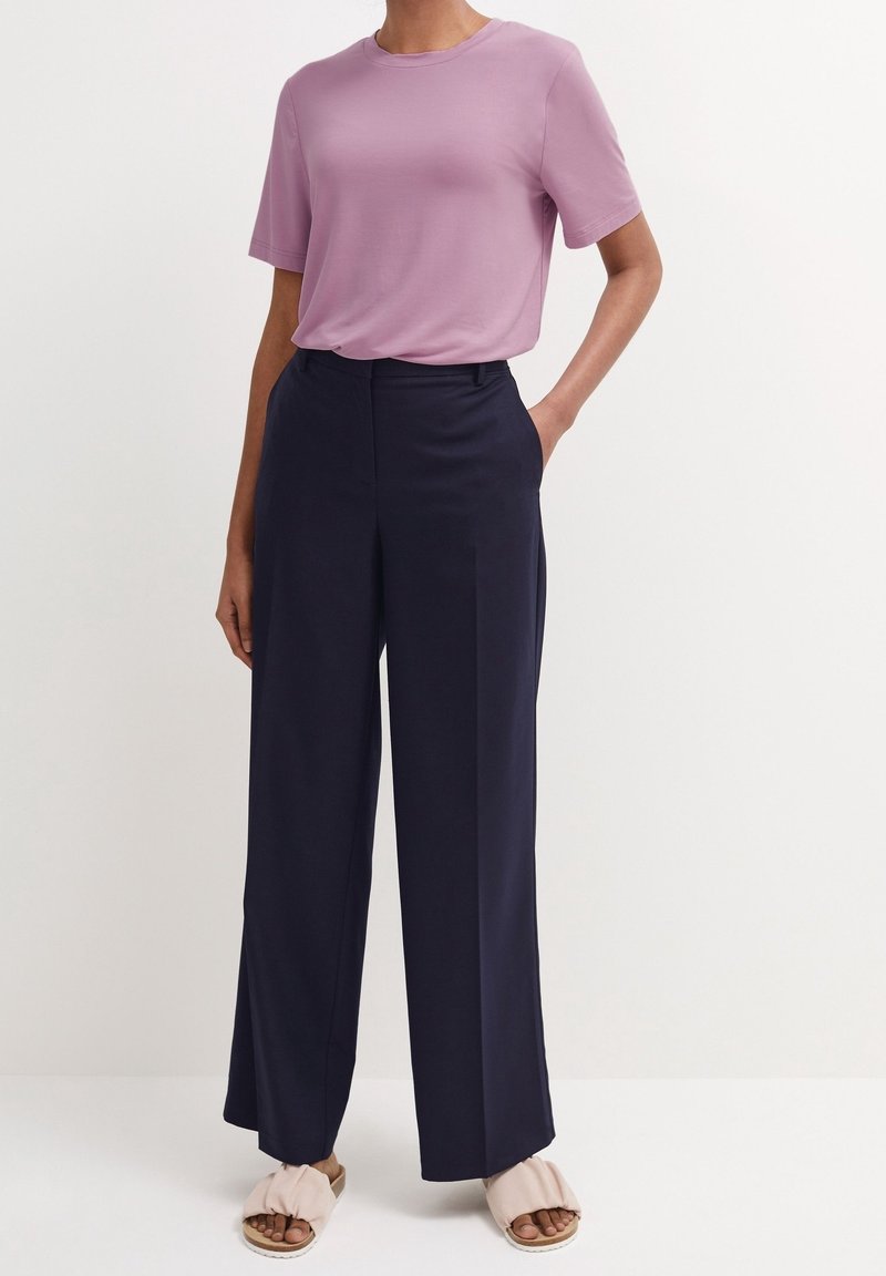 Femme portant un haut violet clair à manches courtes, un pantalon large bleu marine taille haute et des sandales beiges sans bride, debout devant un fond blanc.