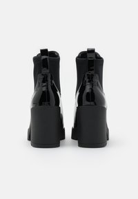 Botas de tobillo de charol negro con tacón grueso, que presentan un panel de tela texturizada y una pestaña de tirón en forma de bucle en la parte posterior.
