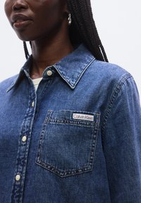 Tummansininen farkkupaita, jossa on terävä kaulus, etutasku ja Calvin Kleinin logo. Pehmeä tekstuuri ja napillinen malli.