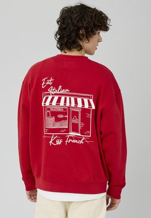 Sweat-shirt rouge présentant une devanture illustrée en blanc avec un auvent rayé, le texte "Eat Italian" et "Kiss French", ainsi qu'une porte détaillée.