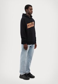 Schwarzer Hoodie mit "ICECREAM" in bunten Buchstaben, kombiniert mit hellblauen Jeans und schwarzen Sportschuhen. Einfaches Design, lockere Passform.