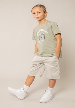 Garçon debout les mains dans les poches, portant un t-shirt vert avec un motif de moto, un short beige, des chaussettes blanches et des baskets montantes blanches.