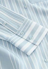 Chemise à manches longues rayée bleu clair et blanc avec poignets boutonnés et boutons sur le devant, montrant la texture du tissu et les détails des coutures.