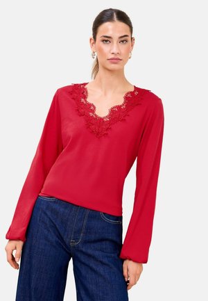 Frau trägt ein rotes Langarmshirt mit floraler Spitzen-V-Ausschnitt-Detail und dunkelblaue Jeans vor neutralem Hintergrund.