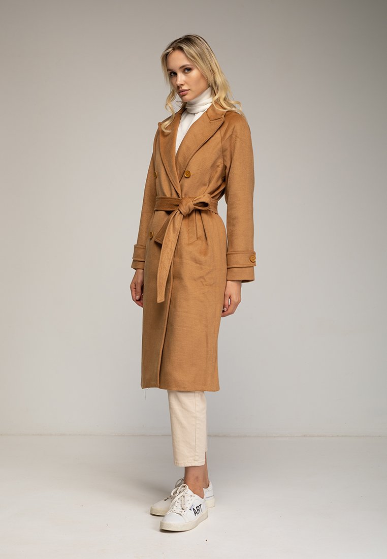 Cappotto avvolgente color cammello con bottoni doppi, cinturino e tasche laterali. Abbinato a un dolcevita crema e sneakers bianche. Sfondo neutro.