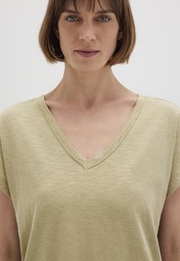 V-neck, short-sleeved top in light olive green, made of soft, textured fabric with subtle variations in color and stitching detail.