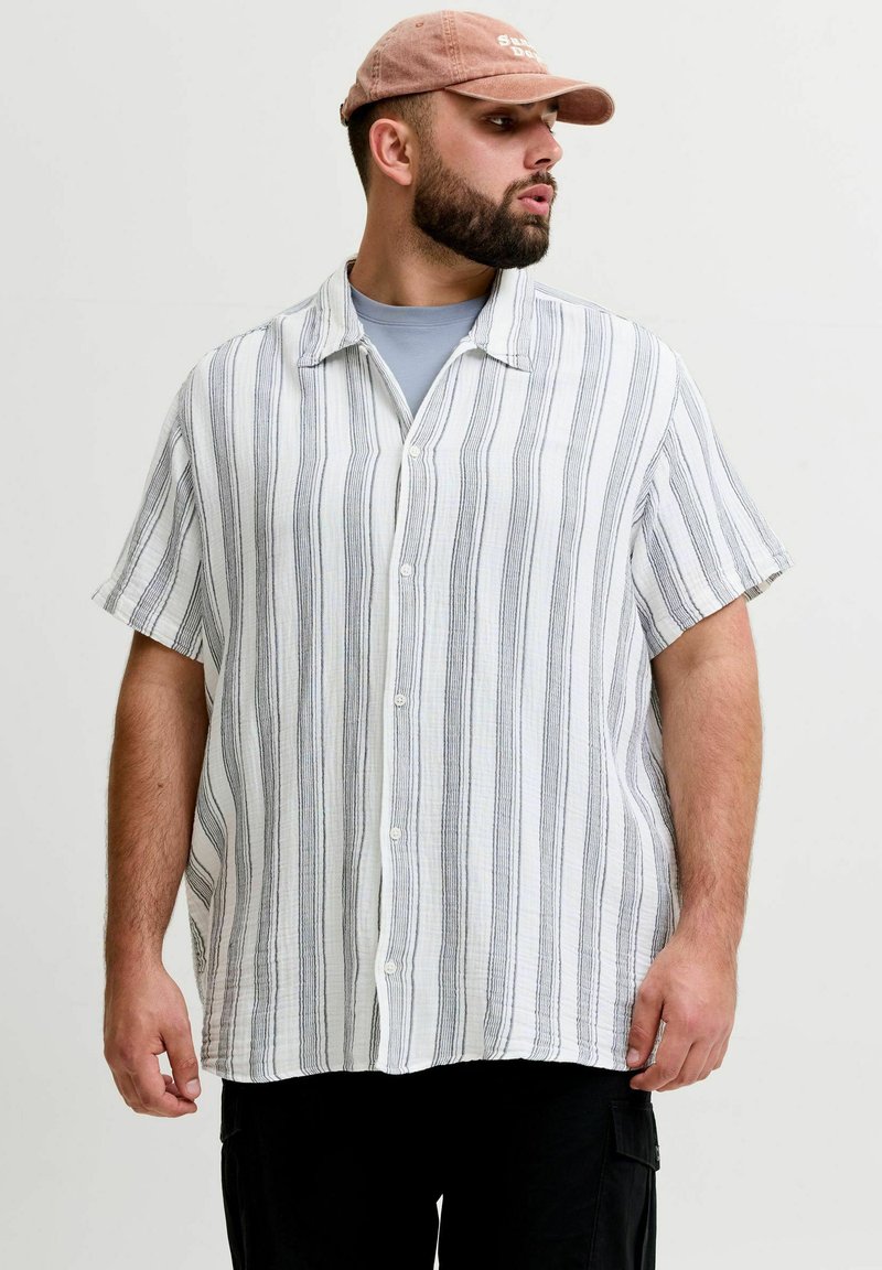 Muž s bradou, ktorý má na hlave ružovú čiapku, bielo-sivú vertikálne pruhovanú krátkorukávovú košeľu cez sivé tričko a čierne nohavice, pozerá sa do strany.