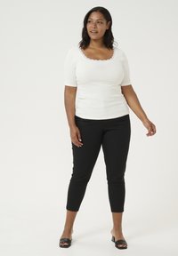 Haut blanc à manches courtes avec un col festonné, associé à un pantalon noir ajusté et des sandales à talons noires, se tenant sur un fond clair.