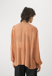 Lett, gjennomsiktig oransje bluse med lange ermer, løs passform og avrundet hem, med subtile folder bak.