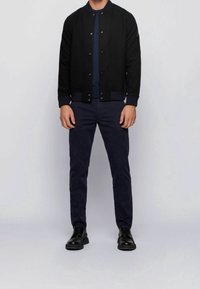 Černá bunda bomber z vlněné směsi s žebrovanými manžetami a spodním lemem, nošená přes námořnický svetr. Spárována s námořnickými kalhotami a černými koženými botami.