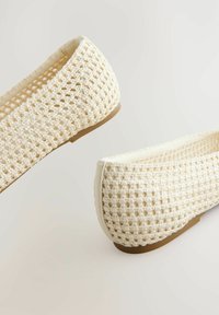 Valkoiset kudotut ballerinat, joissa on teksturoitu, avoin neulekuvio ja litteä, ruskea kumipohja. Pyöristetty kärki ja minimalistinen reunusdetalji.