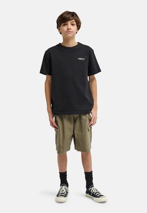 Garçon debout, portant un t-shirt noir avec un petit logo blanc, un short cargo olive, des chaussettes noires et des baskets basses noires sur fond blanc.