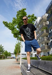 Svart grafisk t-skjorte med vingemønster, denimshorts, hvite sokker og svarte joggesko. Utendørs med trær og moderne bygninger.