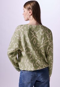 Pitkähihainen vihreä paita, jossa on kermahenkinen abstrakti kuvio, rento leikkaus ja pyöreä kaula-aukko, yhdistettynä sinisiin farkkuihin.