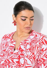 Top de patrón rojo y blanco con escote redondeado, hecho de tela ligera. Combinado con pendientes florales rojos y un maquillaje de ojos rosa sutil.